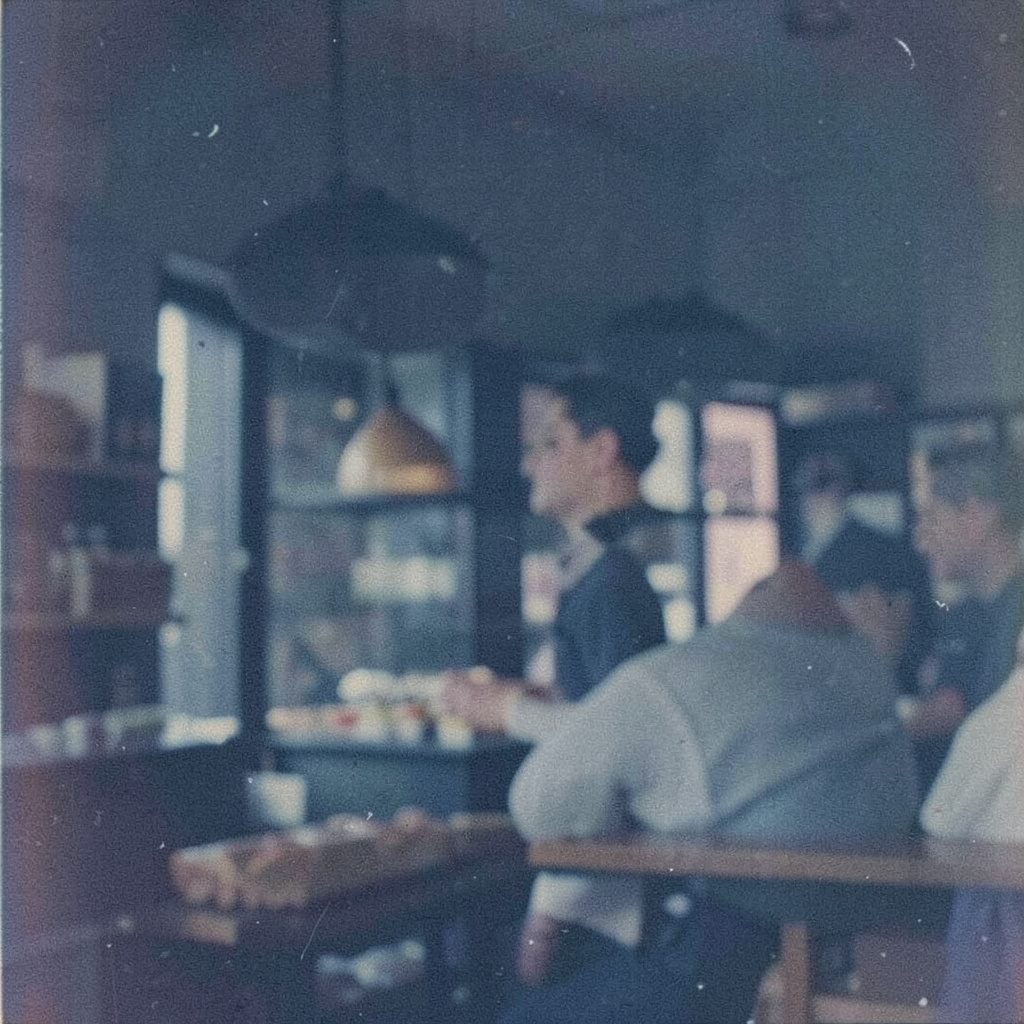 A dimly lit restaurant entrance photographed on grainy film, warm amber light against deep navy shadows, an atmosphere of quiet formality